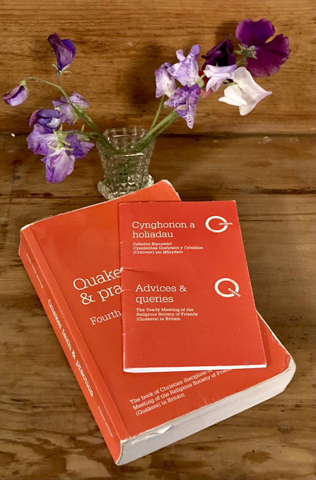 A red book - Quaker Faith & Practice, with a red booklet on top: Cynghorion a holiadau ~ Advices & Queries. On brown well worn wood, with a small glass vase and some mauve and white sweet peas. Both books show signs of wear